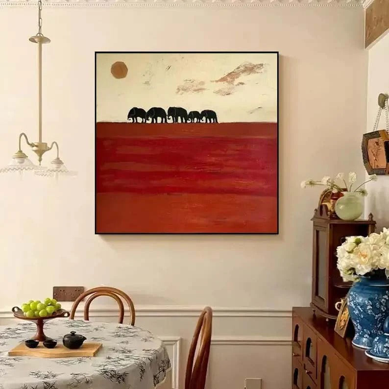a dining room with a large painting of elephants on the wall above a table with chairs. The painting is a vibrant red and white landscape with a clear blue sky. The dining table is set with a blue vase and a white teapot, and there are several chairs around the table.