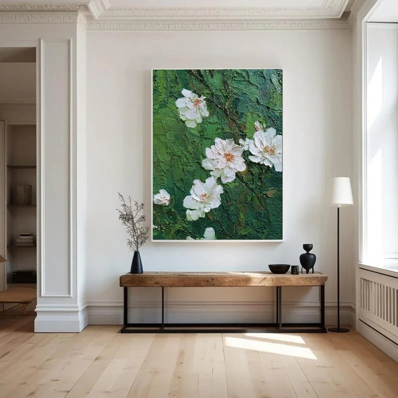a large painting of white flowers on a green background hanging on a white wall in a room with a wooden bench and a black vase.