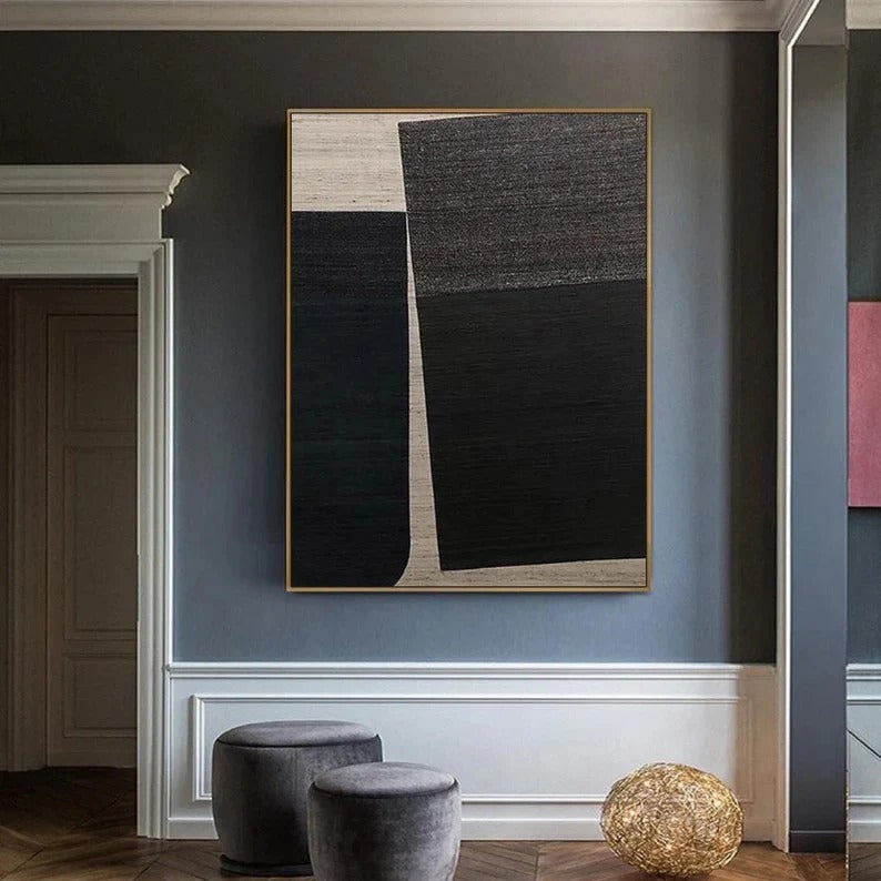 a modern art piece hanging on a gray wall, with a white frame. The art piece is a large abstract painting with black and white colors. In the foreground, there are two gray ottomans placed on a wooden floor.