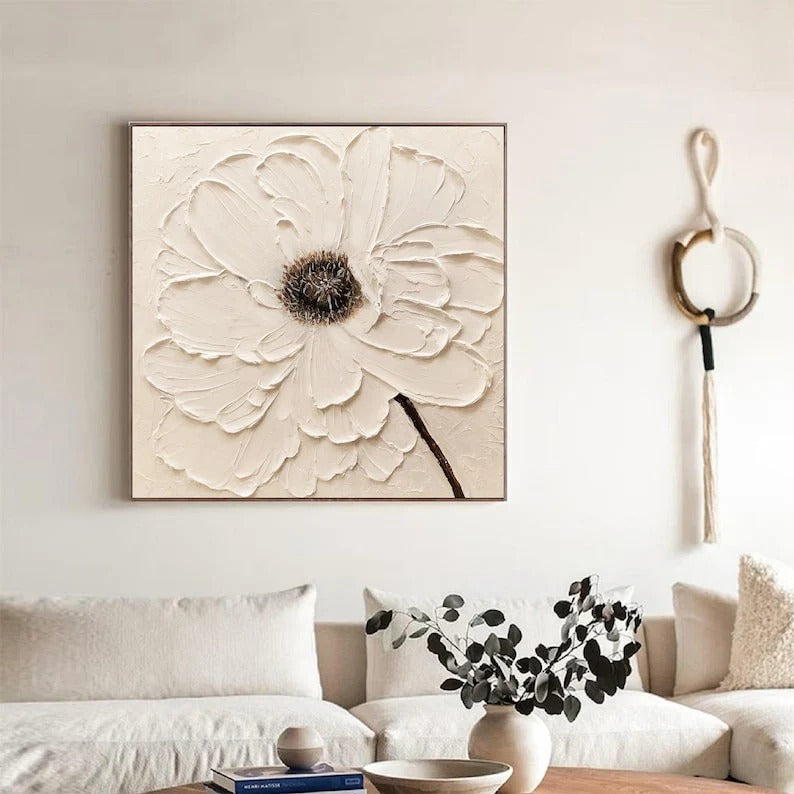 a living room with a large white flower painting hanging on the wall above a couch. The painting is a square canvas with a detailed, textured flower design. The room has a neutral color scheme with white walls and a beige couch. There is also a vase with greenery on the coffee table, adding a touch of nature to the space.
