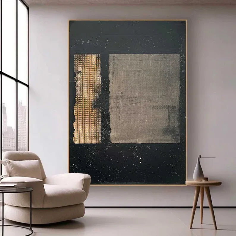 a large abstract painting hanging on a wall in a modern living room. The painting has a black background with a white square and a gold grid pattern on the left side. In the foreground, there is a beige armchair and a small wooden side table with a vase on it.