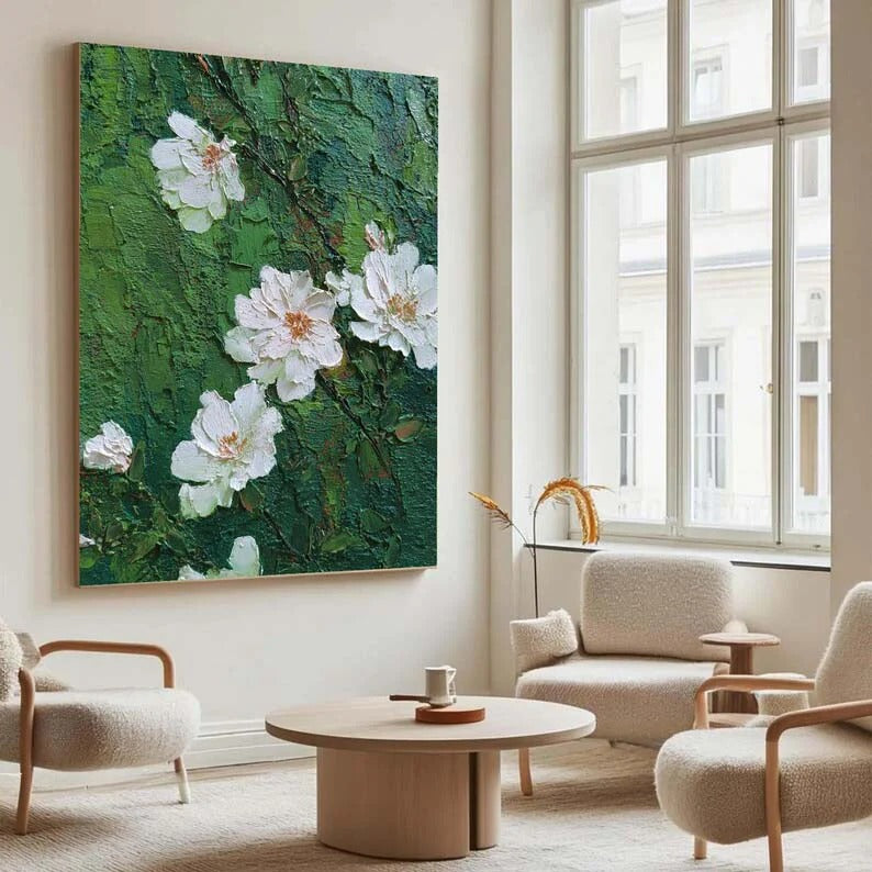 a modern living room with a large painting of white flowers on a green background hanging on the wall. The room is furnished with two beige armchairs and a round wooden coffee table, creating a cozy and inviting atmosphere.