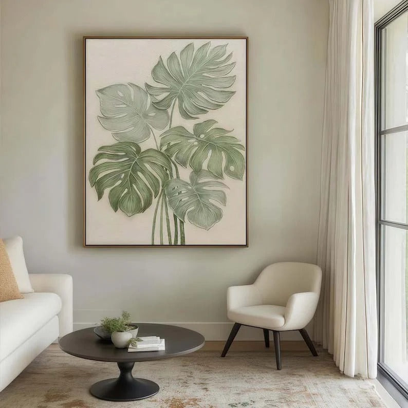 a living room with a large framed painting of green monstera leaves hanging on the wall. The room has a white sofa, a round coffee table, and a white armchair. The overall decor has a modern and minimalist style.