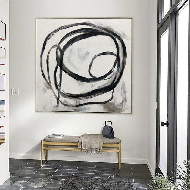 a large abstract painting hanging on a white wall above a wooden bench. The painting has a black and white color scheme with bold, expressive brushstrokes. The bench is positioned in front of the painting, creating a focal point in the room.