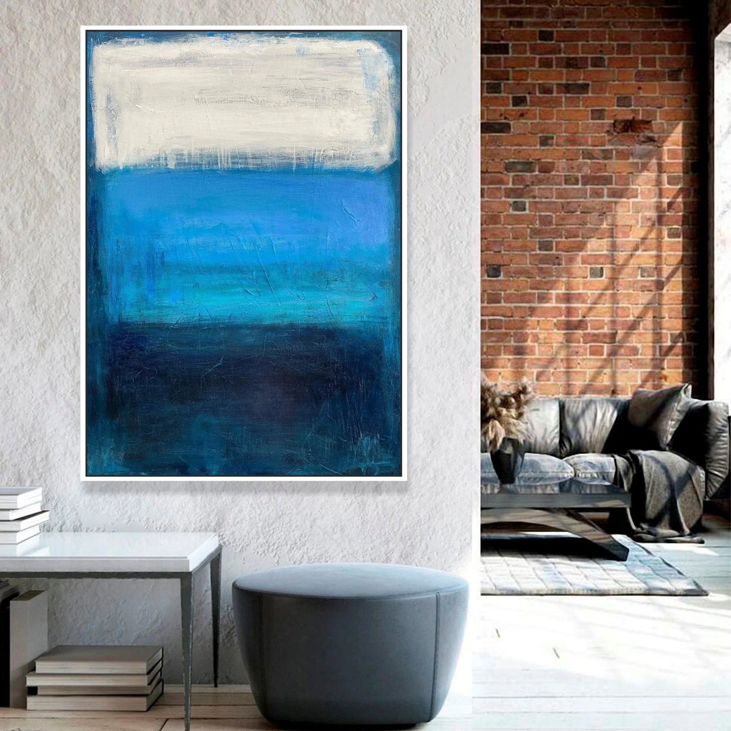 a modern living room with a large abstract painting of blue and white colors hanging on the wall. The painting is framed in white and is positioned above a gray ottoman. In the background, there is a brick wall and a couch, creating a cozy and stylish atmosphere.