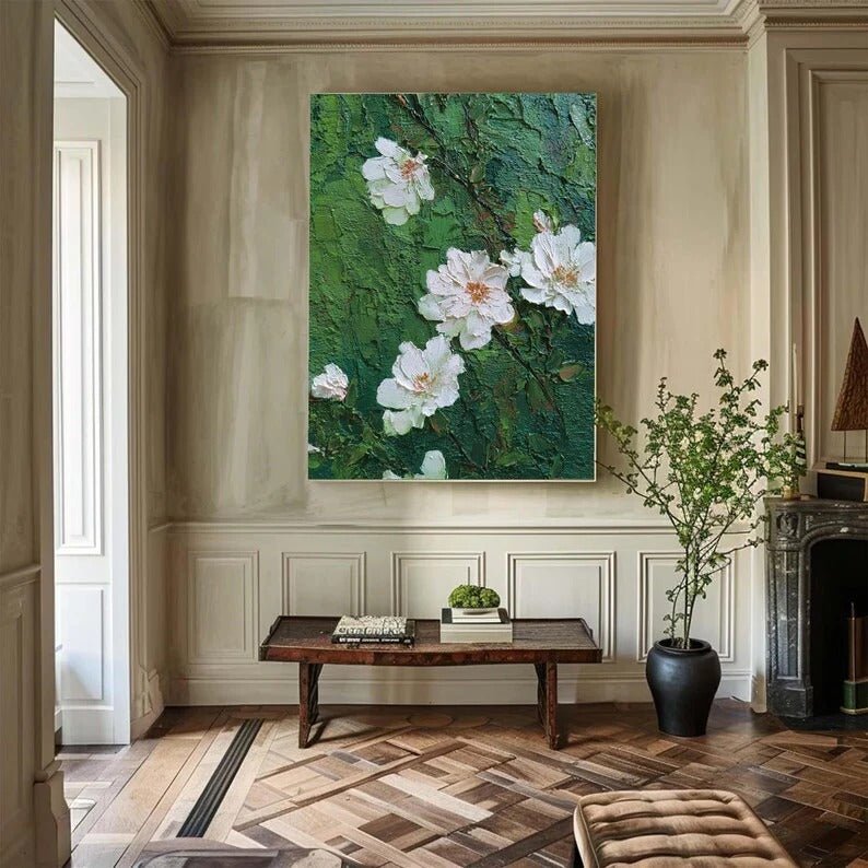 a living room with a large painting of white flowers hanging on the wall above a wooden coffee table. The room has a fireplace, a potted plant, and a wooden floor.