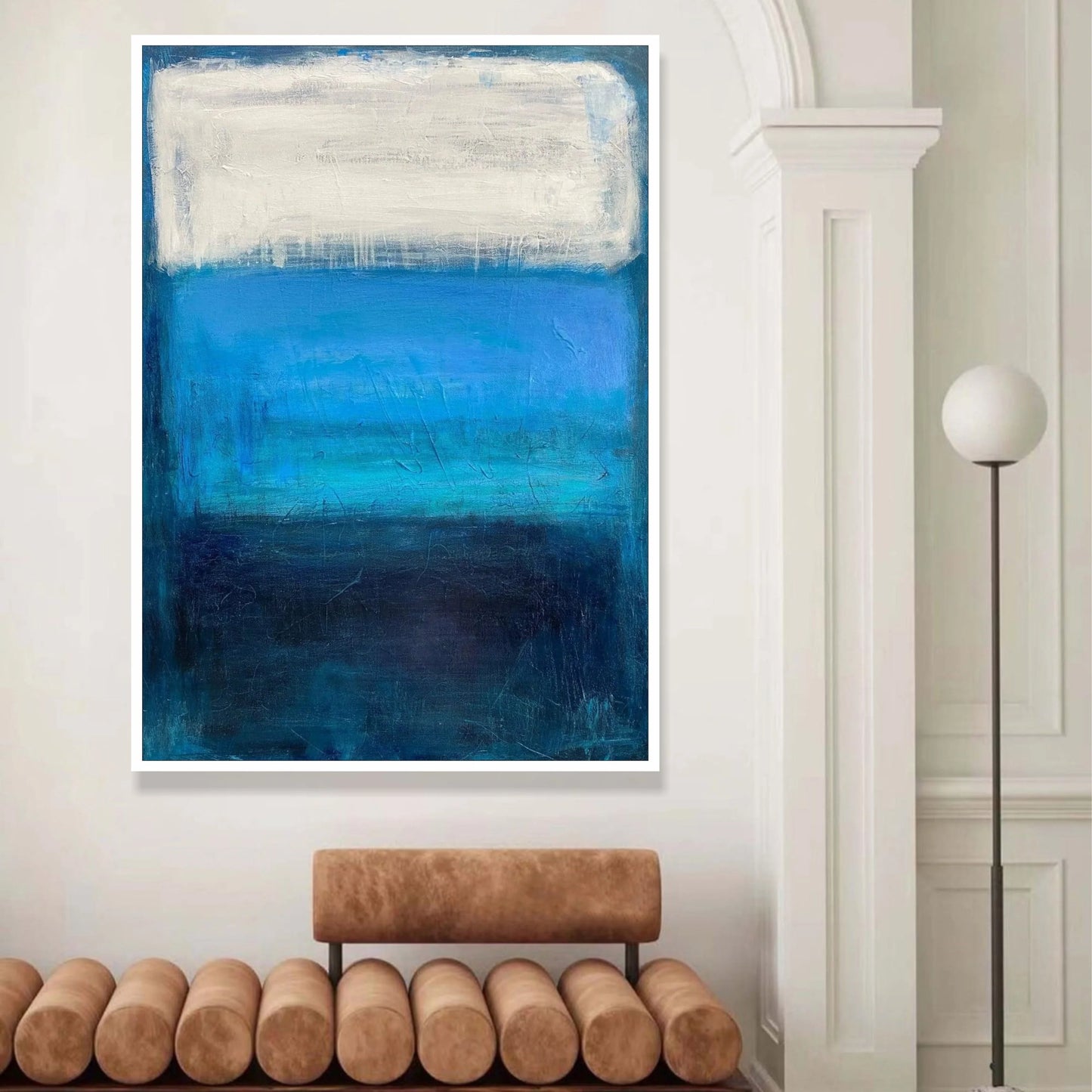 a large abstract painting with bold blue and white colors, hanging on a white wall above a brown bench.