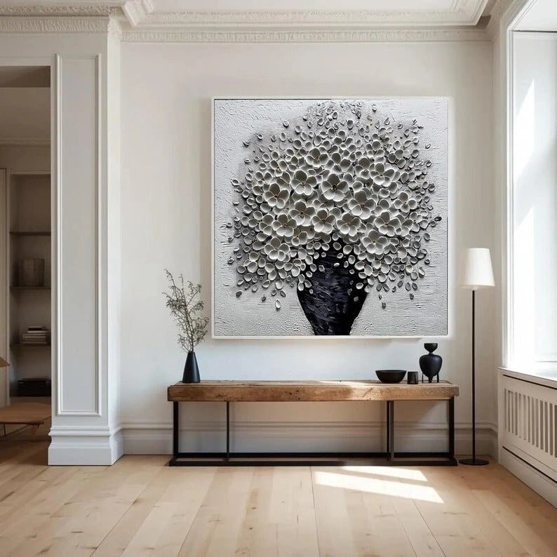 a large, black and white floral painting hanging on a white wall above a wooden console table. The console table has a vase and a bowl on it, and there is a lamp on the right side of the table. The room has a clean, minimalist aesthetic with white walls and a wooden floor.