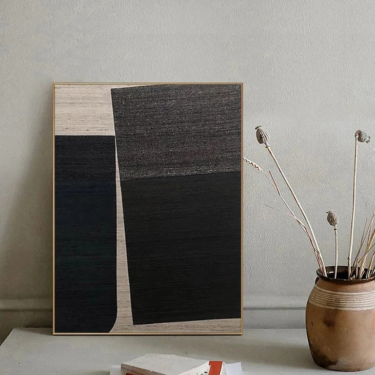 a minimalist art piece with a black and white abstract design, displayed on a wall next to a vase with dried flowers.
