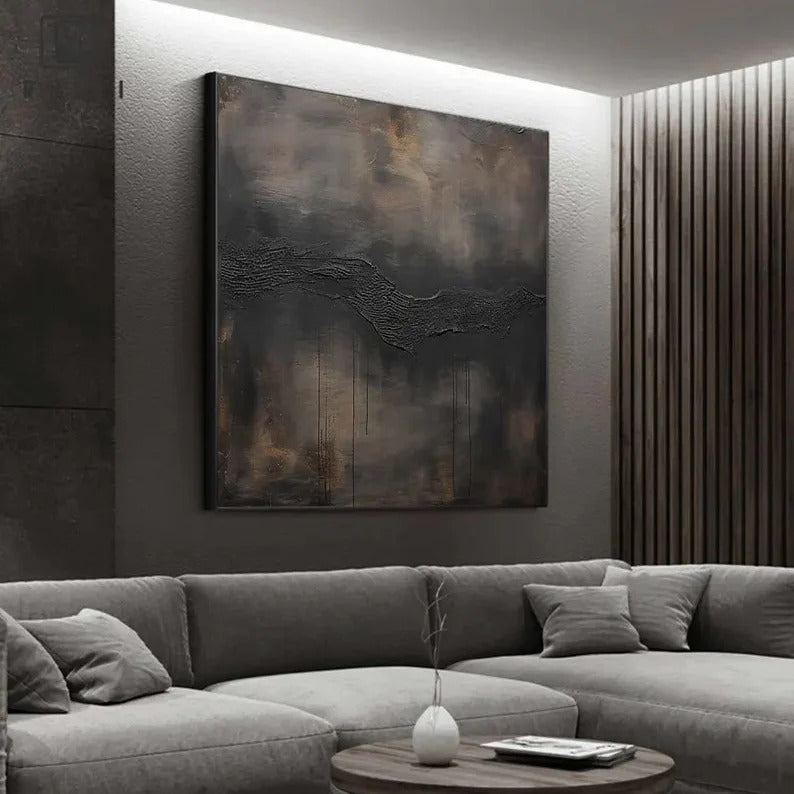 a modern living room with a large abstract painting hanging on the wall above a gray sectional sofa. The painting has a dark, moody color scheme with a textured, black and brown design. The room has a minimalist and contemporary aesthetic, with a wooden floor and a round coffee table in the center.