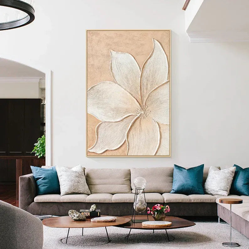 a living room with a large floral painting hanging on the wall above a beige sofa. The sofa is adorned with blue and white throw pillows. In front of the sofa, there is a coffee table with a vase of flowers and a book. The room also includes a dining table with a vase of flowers and a book, creating a cozy and inviting atmosphere.