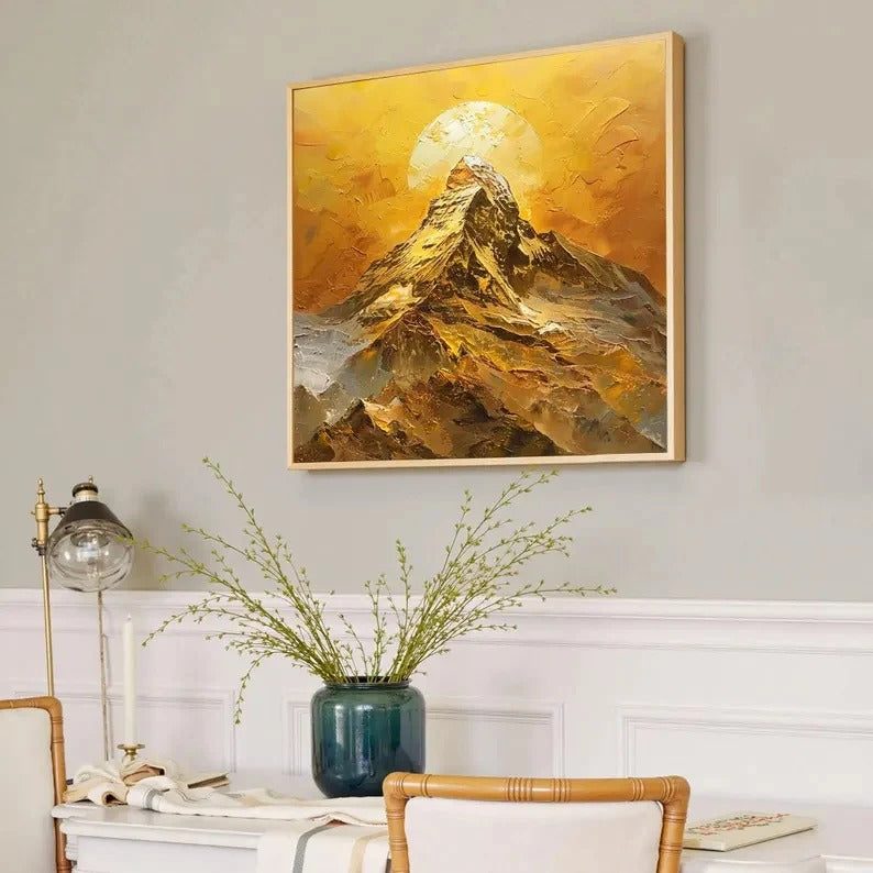 a framed painting of a mountain landscape with a sun in the background, hanging on a wall above a white table with a vase of greenery.