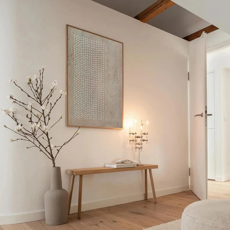 a serene and minimalist living space with a large abstract painting on the wall, a wooden bench, a vase with white flowers, and a lit candle on a table.