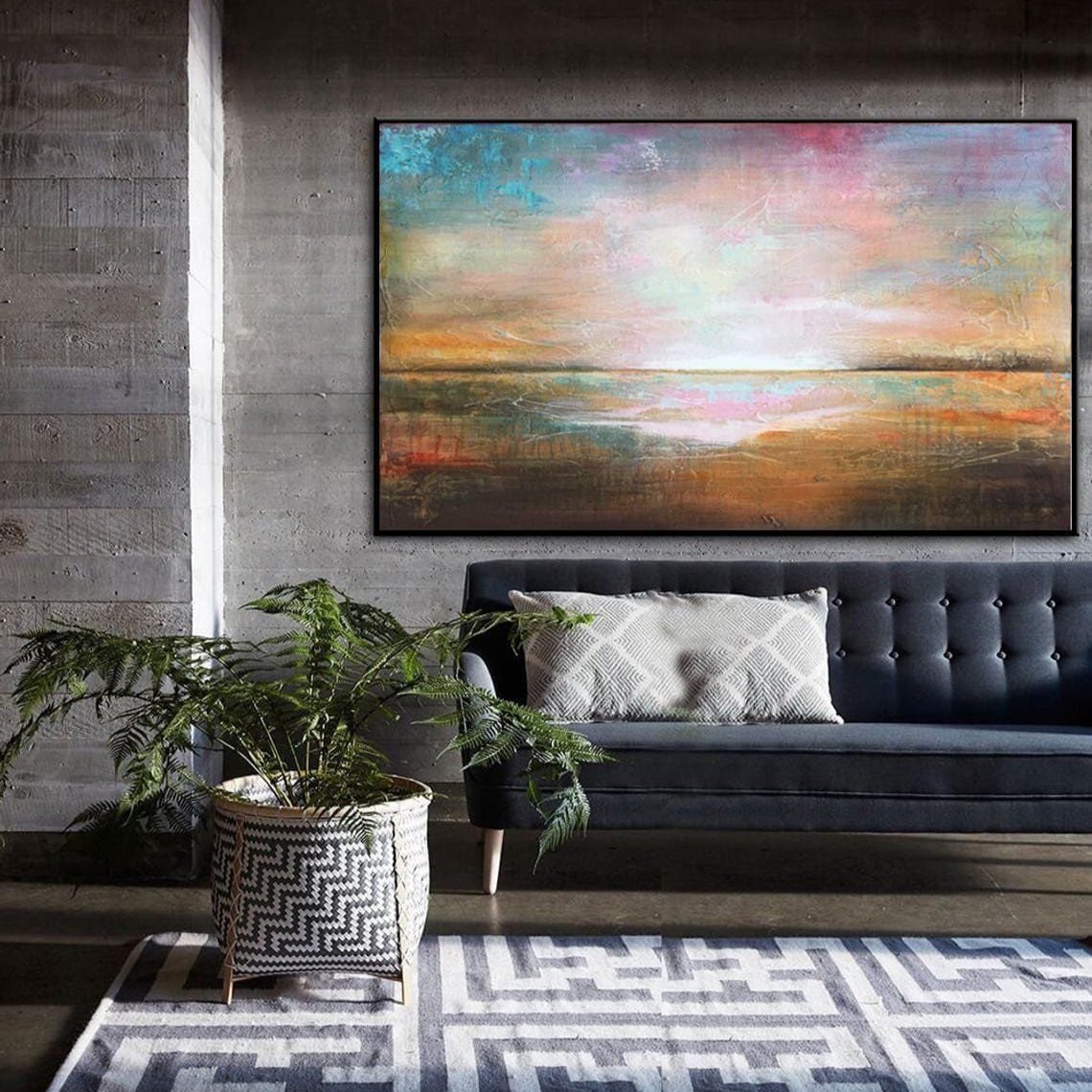 a living room with a large abstract painting hanging on the wall above a black sofa. The painting depicts a colorful landscape with a sunset and mountains in the background. The room also includes a potted plant and a geometric patterned rug, creating a cozy and inviting atmosphere.