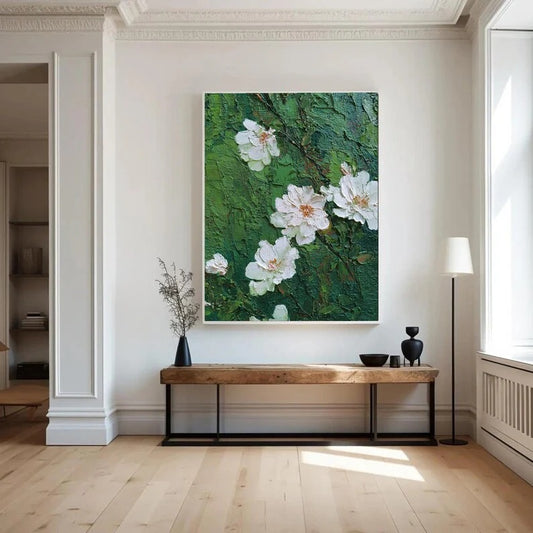 a large painting of white flowers on a green background hanging on a white wall in a room with a wooden bench and a black vase.