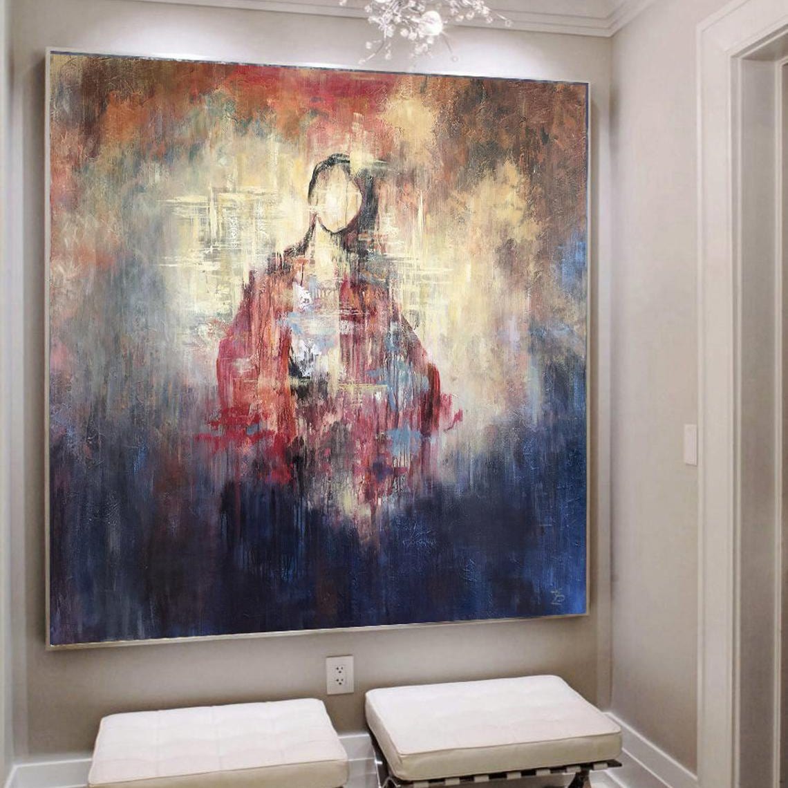 a large, colorful abstract painting hanging on a wall. The painting is predominantly red, blue, and yellow, with a central figure that appears to be a woman. The painting is quite large, covering a significant portion of the wall. In the foreground, there are two white ottomans placed against the wall, providing a contrast to the vibrant colors of the painting.