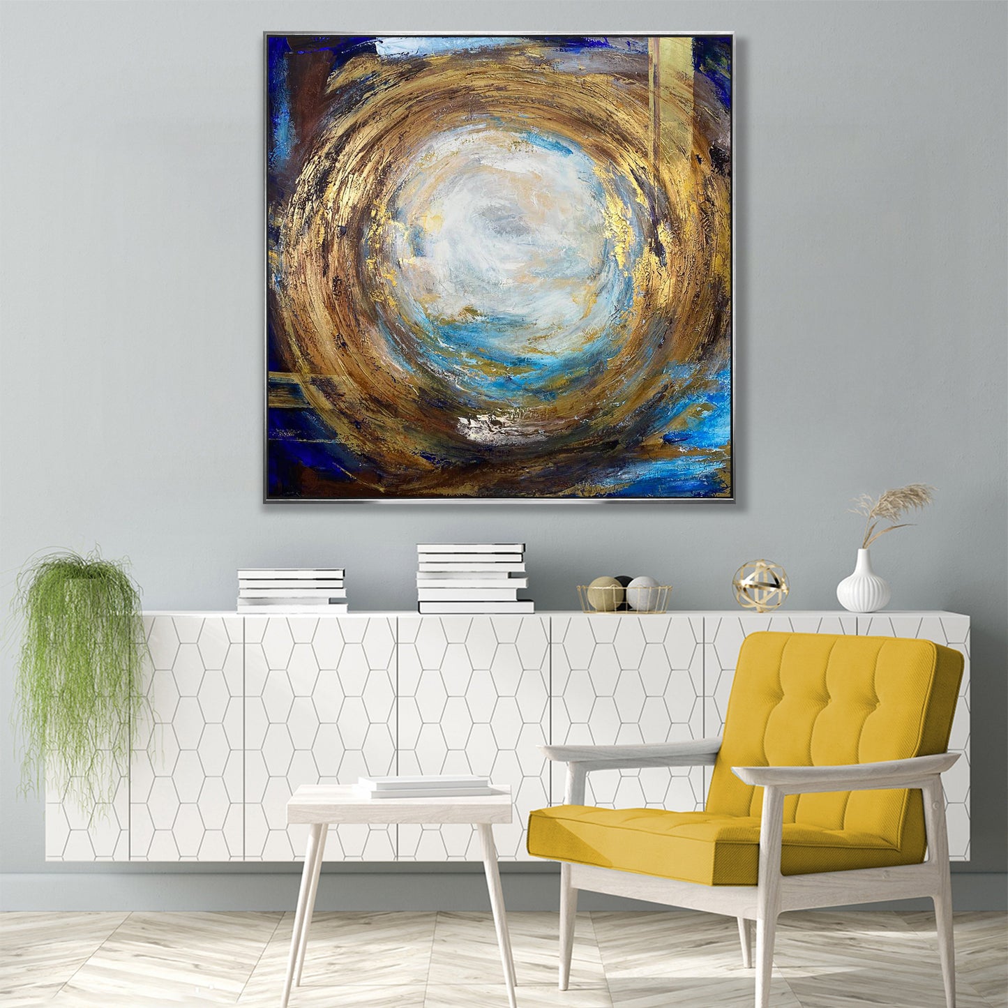 a modern living room with a large abstract painting hanging on the wall above a white cabinet. The painting has a circular design with a mix of blue, gold, and white colors. The room also includes a yellow armchair, a white side table, and a potted plant.