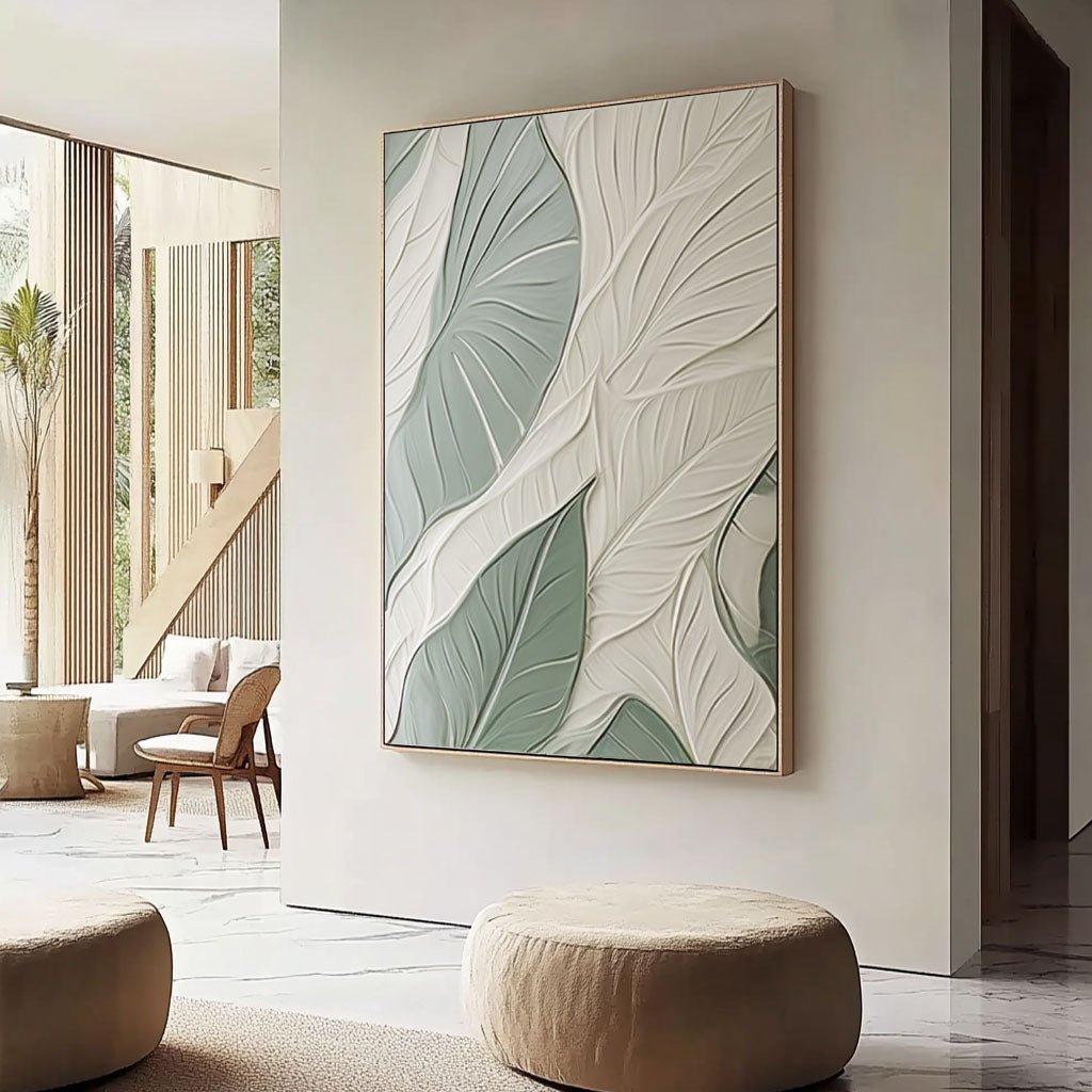 a modern living room with a large wall art piece depicting a leaf pattern in shades of green and white. The room is furnished with a sofa, a chair, and a round ottoman, creating a comfortable and inviting atmosphere.
