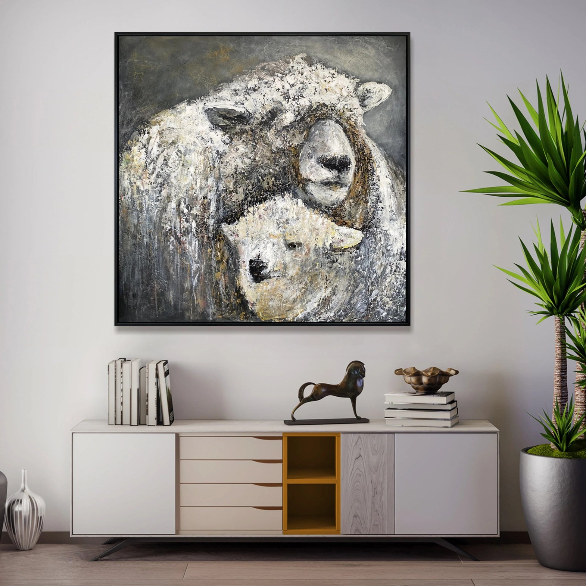 a large painting of two sheep, one white and one brown, hanging on a wall above a white cabinet with drawers. The cabinet also has a small figurine of a horse on top of it. The wall behind the cabinet has a plant, and there are several books and decorative items on the cabinet.