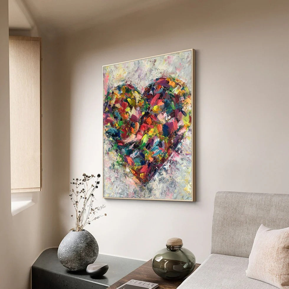 a vibrant, abstract painting of a heart in a gallery setting. The painting is hung on a wall above a wooden table, which has a vase with dried flowers and a small glass jar on it. The overall scene has a cozy and inviting atmosphere, with the heart painting serving as a focal point in the space.