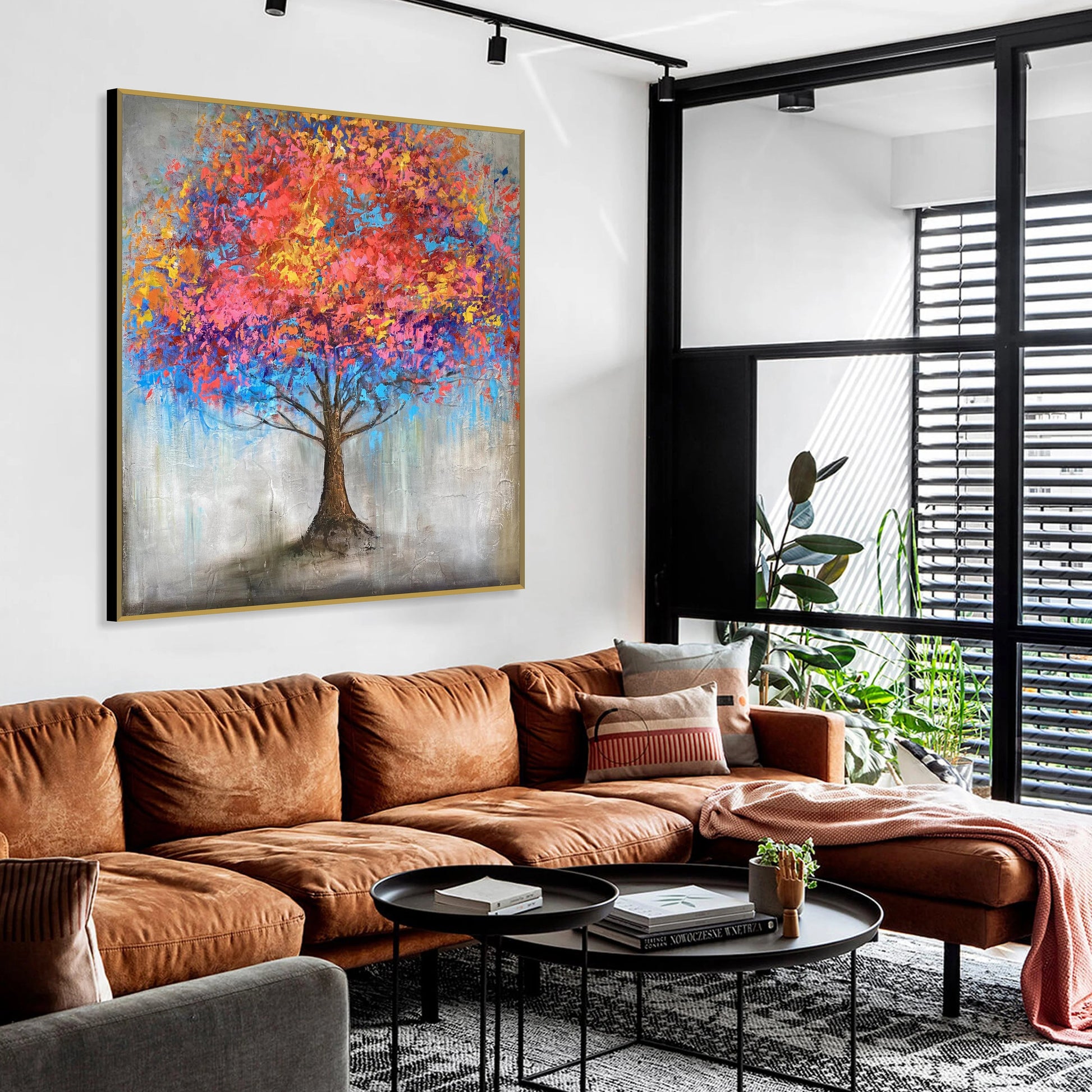 a modern living room with a large, colorful painting of a tree hanging on the wall. The room has a brown leather sofa, a black coffee table, and a large window with blinds.