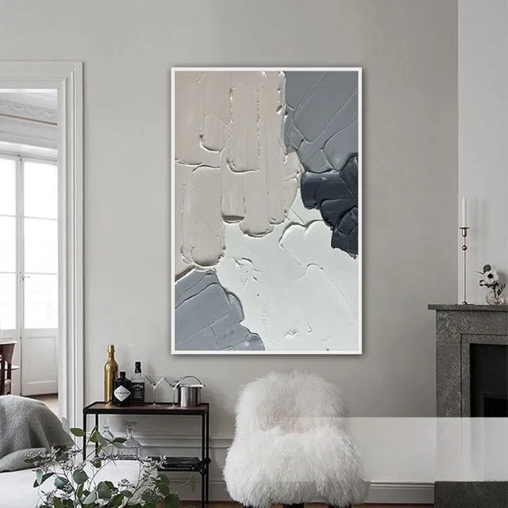 a living room with a large abstract painting hanging on the wall. The painting has a mix of gray and white colors, creating a visually striking contrast. The room also includes a fireplace, a small table with a plant, and a plush white chair.