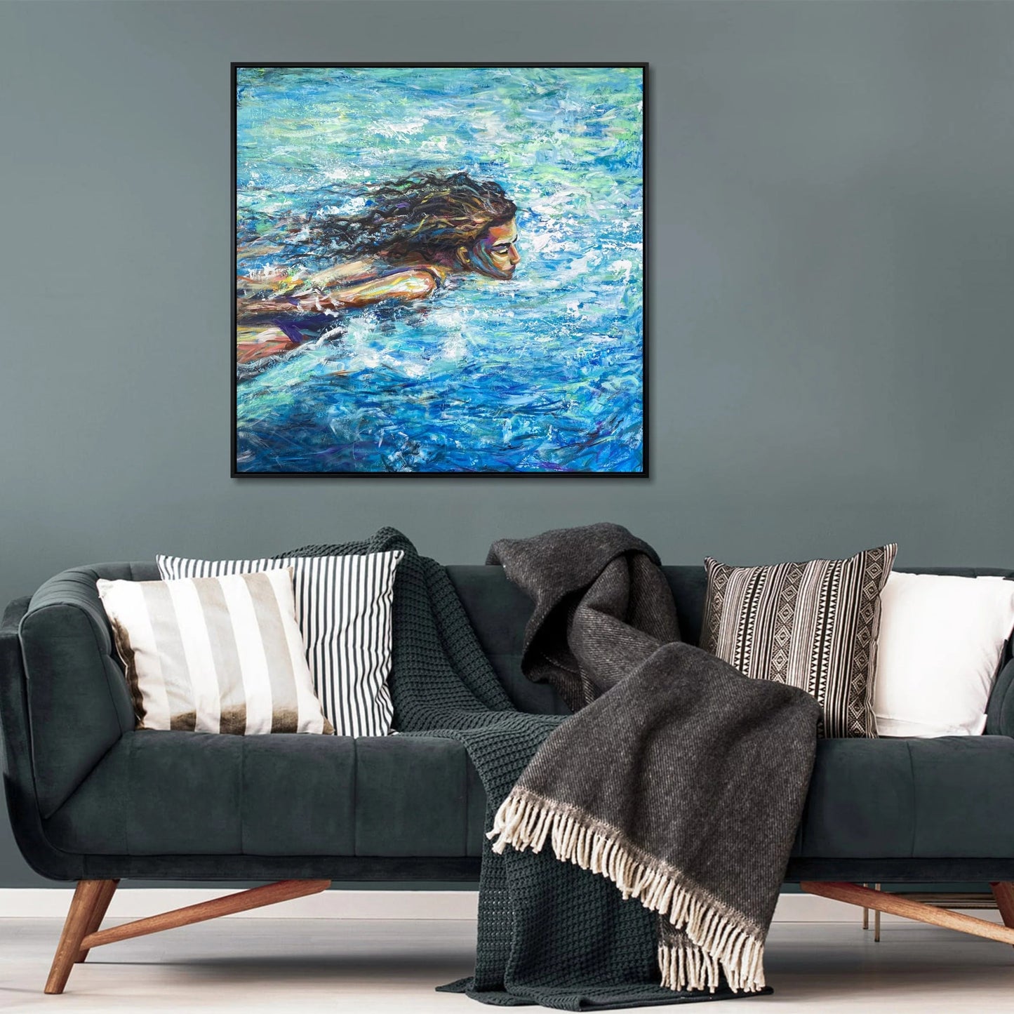 a large painting of a woman swimming in a blue ocean, hanging on a wall above a couch with striped pillows and a gray blanket.