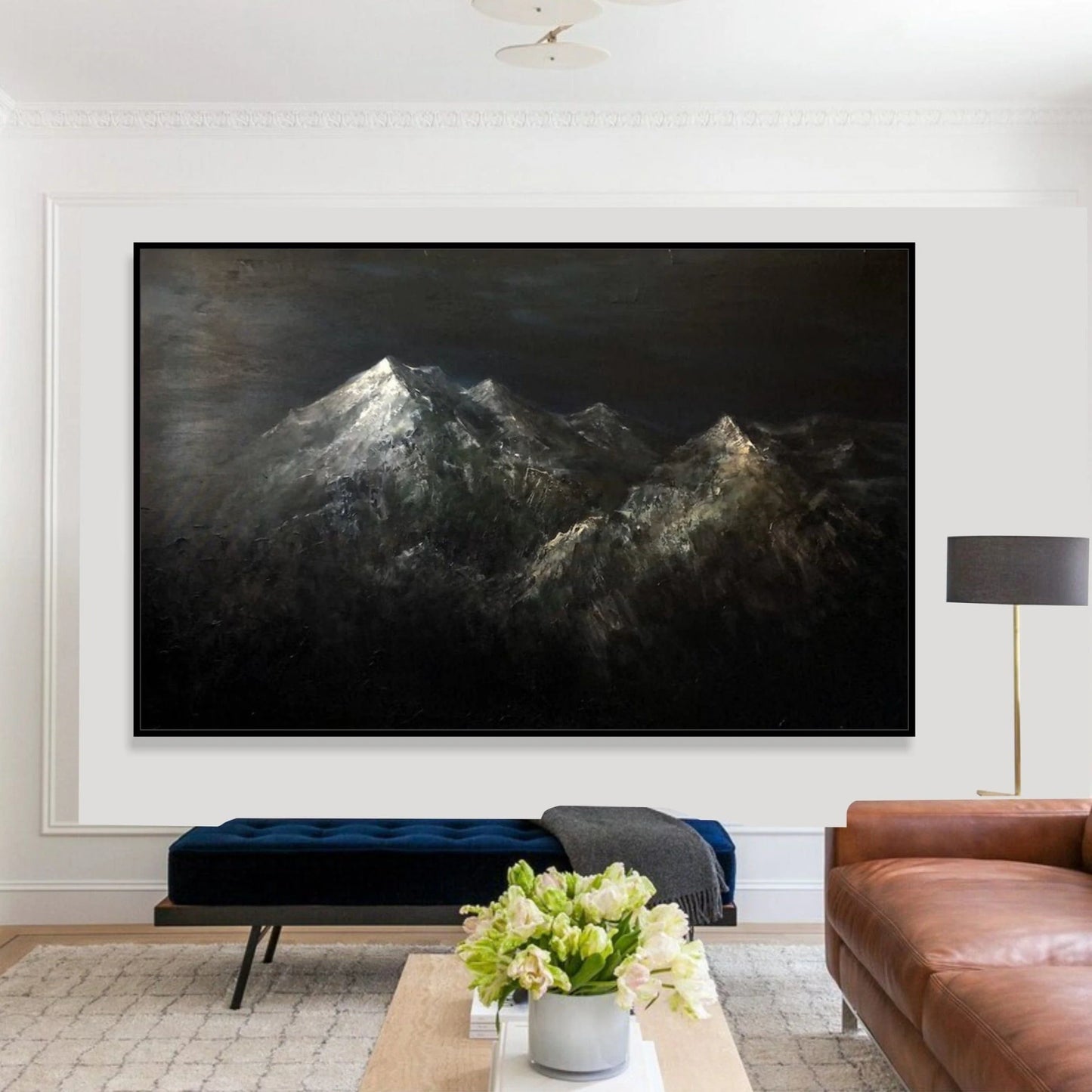 a large, dark-toned painting of a mountain range hanging on a white wall in a living room. The painting is framed and appears to be the focal point of the room. In front of the painting, there is a brown leather couch and a wooden coffee table with a vase of flowers on it.