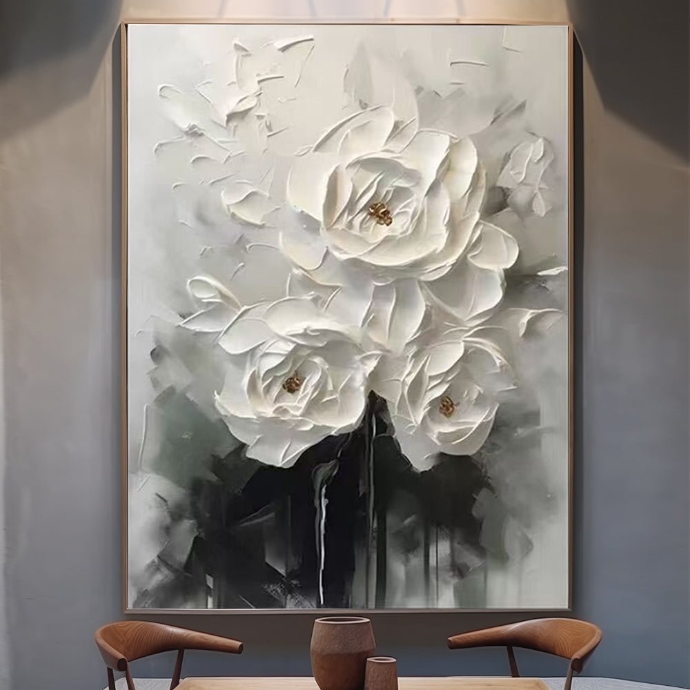 a large, framed painting of white flowers with gold accents, displayed on a wall. In front of the painting, there is a wooden table with two chairs, creating a cozy and inviting atmosphere.