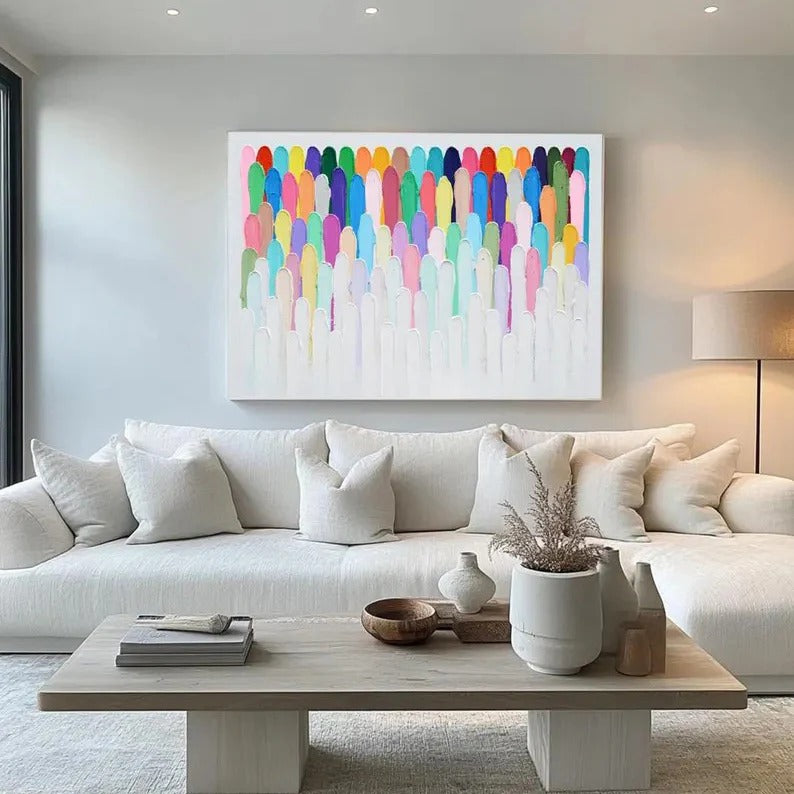 a modern living room with a large, colorful abstract painting hanging on the wall above a white sofa. The sofa is adorned with white pillows and a wooden coffee table in front of it. The coffee table has a vase, a bowl, and a book on it, creating a cozy and inviting atmosphere.