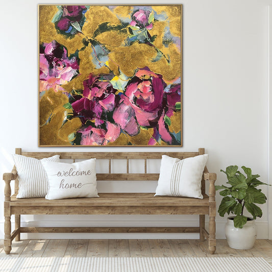 a large, colorful painting of flowers hanging on a wall above a wooden bench with two white pillows. The painting is vibrant with shades of pink, purple, and yellow, creating a warm and inviting atmosphere. The bench is positioned in front of a white wall, and a potted plant is placed next to it, adding a touch of greenery to the scene.