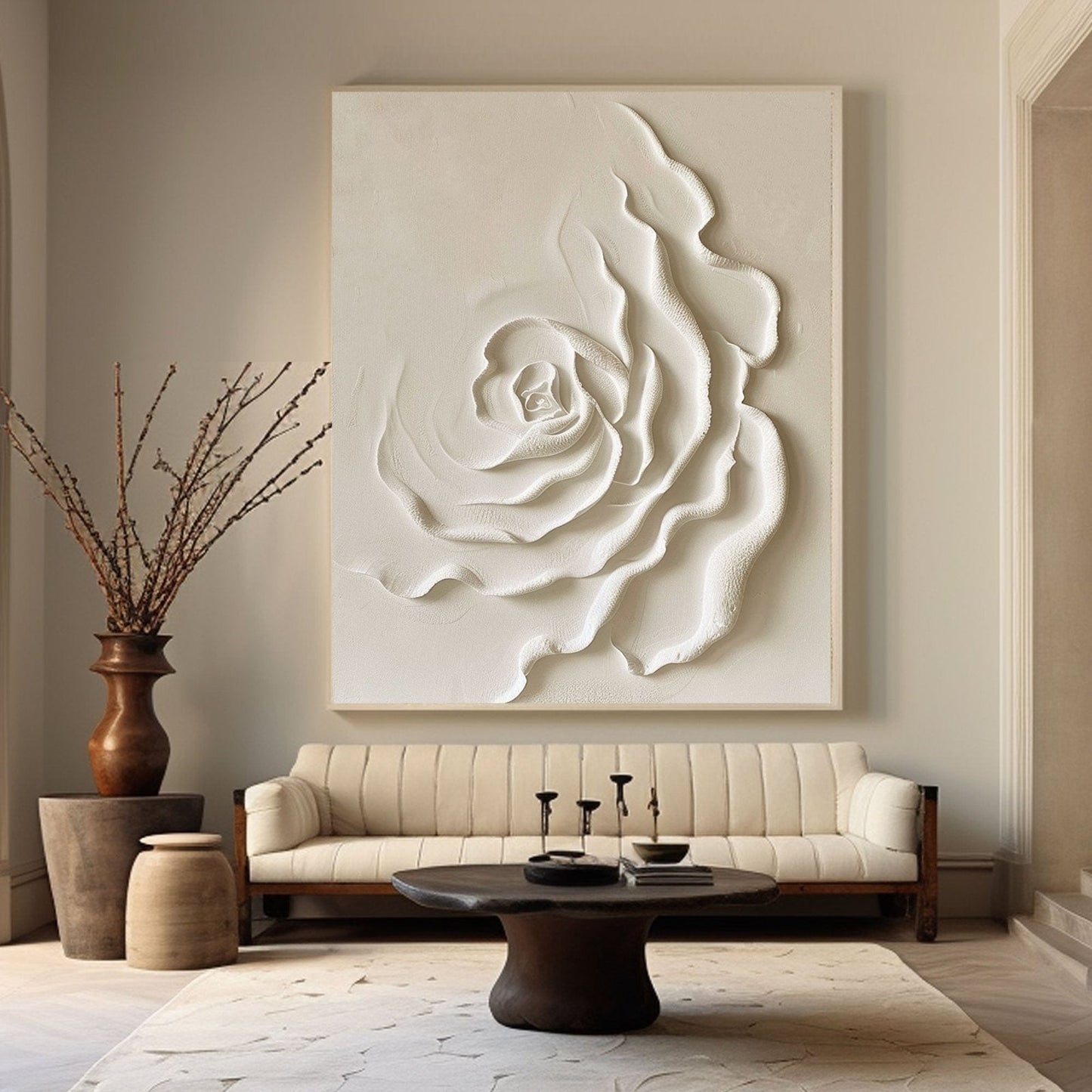 a living room with a large white floral artwork hanging on the wall above a beige sofa. The sofa is positioned in front of a coffee table with a vase of branches and a book on it. The room has a neutral color scheme with a wooden floor and a rug on the floor.
