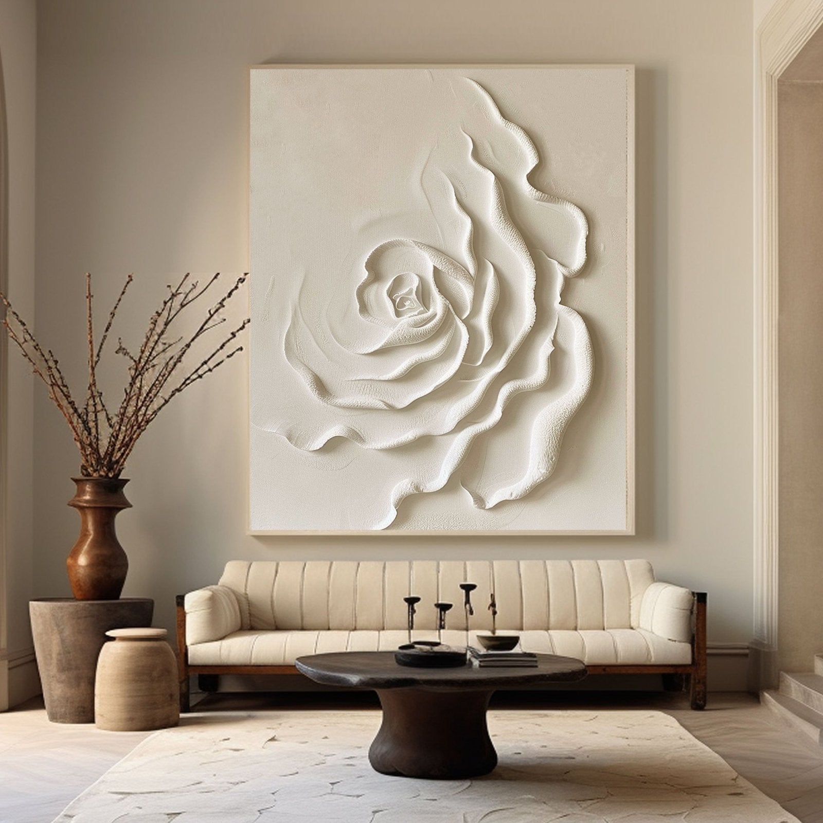 a living room with a large white floral artwork hanging on the wall above a beige sofa. The sofa is positioned in front of a coffee table with a vase of branches and a book on it. The room has a neutral color scheme with a wooden floor and a rug on the floor.