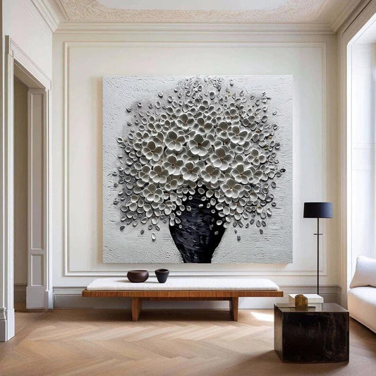 a large, white, floral-patterned painting hanging on a wall in a modern living room. The painting is mounted above a wooden bench, which has two small bowls placed on it. The room also includes a black side table with a lamp and a white sofa.