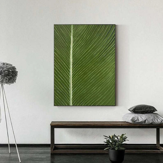 a large green leaf with a white line running down its center, hanging on a white wall above a wooden bench. The bench is positioned in front of a potted plant, and there are two gray pillows on it.