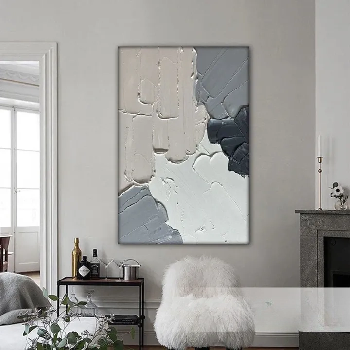 a living room with a large abstract painting hanging on the wall. The painting has a mix of gray and white colors, creating a modern and sophisticated look. The room is furnished with a white chair, a small table with a bottle and a vase, and a fireplace. The overall atmosphere of the room is calm and elegant.