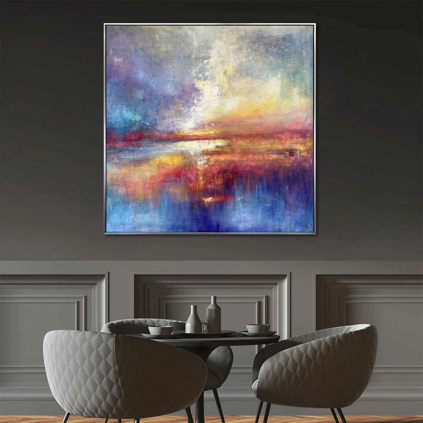 a large abstract painting hanging on a wall above a dining table with four chairs. The painting has a vibrant and colorful composition, with a mix of blue, orange, and purple hues. The dining table is set with a vase, a cup, and a bowl, creating a cozy and inviting atmosphere.