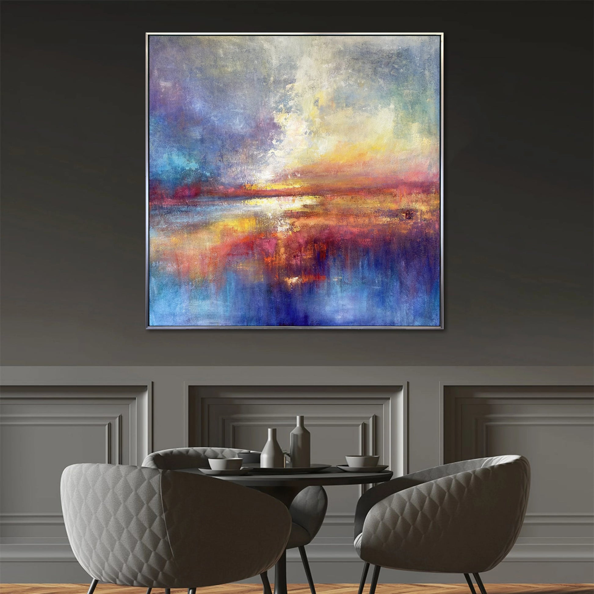 a large abstract painting hanging on a wall above a dining table with four chairs. The painting has a vibrant and colorful composition, with a mix of blue, orange, and purple hues. The dining table is set with a vase, a cup, and a bowl, creating a cozy and inviting atmosphere.