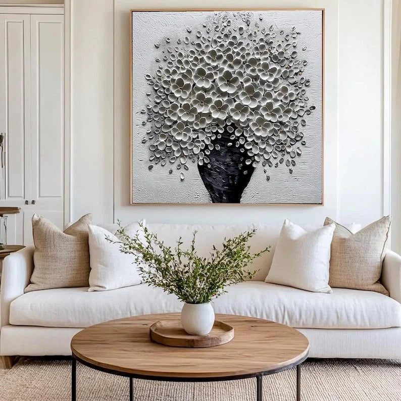 a living room with a large black and white floral painting hanging on the wall above a white sofa. The sofa is adorned with two beige throw pillows. In front of the sofa, there is a wooden coffee table with a white vase containing greenery.