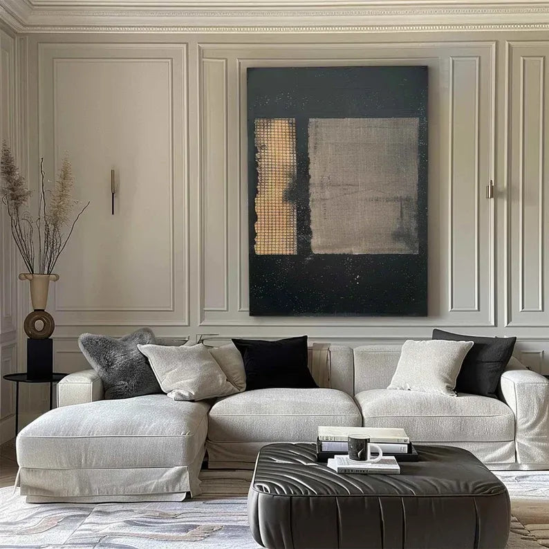 a modern living room with a large abstract painting on the wall, a white sofa with black and gray throw pillows, and a black leather ottoman in front of the sofa.