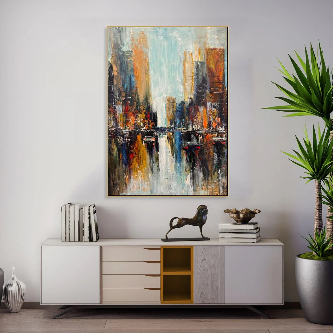 a modern art painting of a cityscape, hanging on a wall above a white cabinet with drawers. The painting is vibrant and colorful, depicting a bustling urban scene with tall buildings and a river flowing through the city. The cabinet below the painting has a decorative metal sculpture of a horse on top, adding a unique touch to the room's decor.