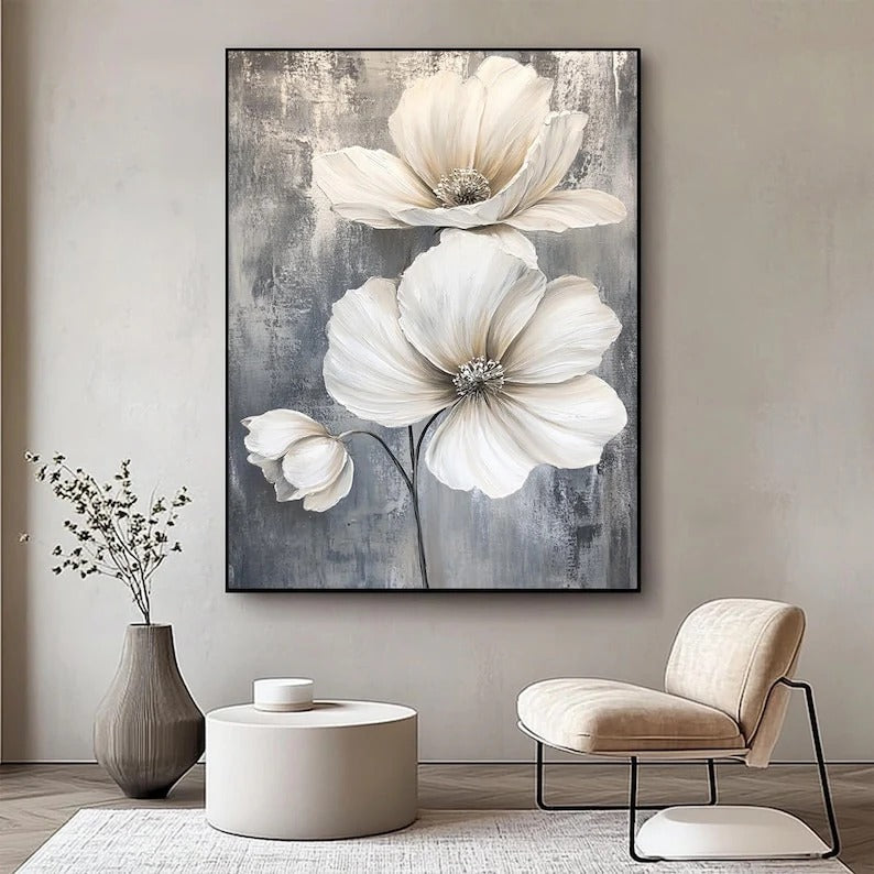 a large painting of white flowers hanging on a wall, with a gray and white color scheme. The painting is positioned above a round coffee table and a beige armchair, creating a cozy and inviting atmosphere in the room.